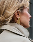 A close-up side profile of a woman with blonde hair wearing a chunky, polished gold geometric hoop earring and a light beige trench coat.