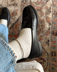 A person wearing black leather penny loafers with off-white ribbed socks and light blue jeans, standing on a patterned area rug. The brand name 'rina_celeste' is watermarked at the bottom.