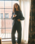 Woman in dark outfit holding a cup by a window with curtains