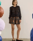 A woman stands in a studio wearing a black long-sleeve lace-trim top and a black shorts with a brown paisley pattern. She is framed by colorful hanging paper lanterns on both sides.