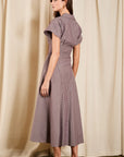 A woman modeling a short-sleeve midi dress with vertical purple and beige stripes, shown from a side-back angle against a draped beige curtain backdrop.