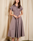 A woman modeling a short-sleeve button-front midi dress with vertical stripes in muted purple and beige, standing against a draped beige curtain backdrop.