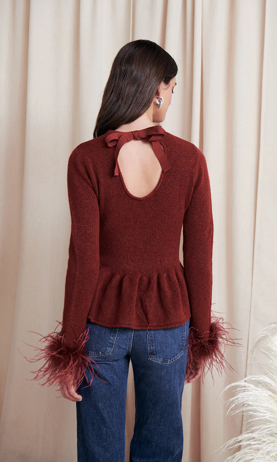 Woman wearing a burgundy sweater with feather details and blue jeans against a beige curtain background, back