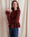 Woman wearing a burgundy sweater with feather details and blue jeans against a beige curtain background, side/front view