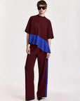 A model wearing a color-blocked two-piece outfit consisting of a short-sleeve top and matching wide-leg trousers. The top is burgundy with a bright blue ruffled overlay, and the trousers are burgundy with a matching blue side stripe.
