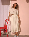 Woman in a white outfit standing next to a red chair against a pink wall.