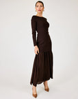 A woman modeling a dark brown long-sleeve maxi dress with a boat neckline and a pleated skirt, standing against a plain light gray studio background.