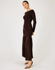 A woman modeling a dark brown long-sleeve maxi dress with a boat neckline and a pleated, flowing skirt, paired with brown pointed-toe heels against a light gray studio background.