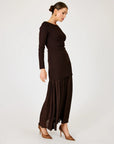 A woman modeling a dark brown ribbed knit long-sleeve maxi dress with a pleated, flowing skirt, paired with brown pointed-toe heels, against a plain light gray studio background.