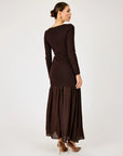 A woman modeling a dark brown long-sleeve maxi dress from the back, featuring a structured waist and a flowing pleated skirt, set against a plain light gray studio background.