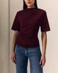 model wearing a short sleeve burgundy top with ruching across the shoulder and jeans on a cream background
