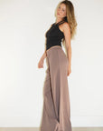 A woman modeling a black halter top and high-waisted, wide-leg taupe maxi pants against a plain white wall and light wood floor.
