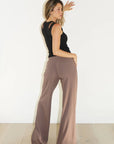 A woman wearing high-waisted, wide-leg taupe pants and a black sleeveless top, standing in profile against a white wall with light wood flooring.