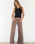 A woman modeling a pair of wide-leg taupe pants with a black sleeveless crop top, standing against a white wall on a light wood floor.