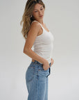 Woman wearing a white tank top and blue jeans against a plain background side view