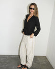 A woman modeling a black long-sleeve wrap-style top and cream wide-leg pants with a thin gold zipper detail, standing in a minimalist room with a white wall and gray concrete floor.