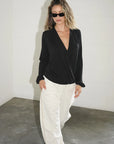 A woman modeling a black long-sleeve wrap-style blouse with a deep V-neck and ruffled cuffs, paired with white wide-leg pants, in a minimalist studio setting with a white brick wall and concrete floor.