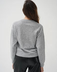 A woman viewed from behind wearing a light gray heathered long-sleeve knit top and black pants, standing against a plain light gray studio background.