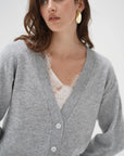 A woman wearing a light gray knit cardigan over a white lace top, photographed against a clean light gray studio background.