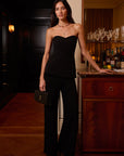 A woman stands in a dimly lit indoor setting wearing a black strapless peplum top and matching wide-leg trousers, holding a small black clutch. A wooden cabinet with bottles and glasses is visible behind her.