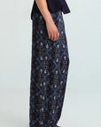 A model is shown from the waist down wearing wide-leg trousers with a detailed blue, black, and white architectural pattern, paired with a dark navy top and brown shoes, against a light gray studio background.