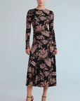 A woman modeling a long-sleeve midi dress with a black base and a dusty rose floral and paisley pattern, paired with black pointed-toe heels against a light gray studio background.