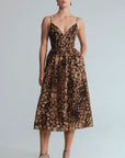 A woman models a brown and beige animal-print midi dress with spaghetti straps and a V-neckline, paired with metallic silver high-heeled sandals, against a light gray studio background.