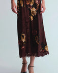 A dark brown midi skirt with a large floral print in beige, gold, and red, featuring a lace hem, shown on a model wearing black pointed-toe heels against a light gray background.
