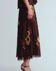 A model wearing a dark brown midi skirt with a gold and orange floral print and a lace hem, paired with black high heels, against a light gray background.