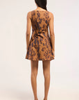 A woman viewed from the back wearing a sleeveless brown and black leopard-print mini dress with a flared skirt, paired with beige heeled sandals, against a plain white studio background.
