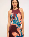 A woman modeling a sleeveless halter-neck dress with a dark burgundy base and a large abstract floral print in teal blue and peach-pink, set against a plain light gray studio background.
