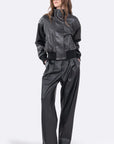 A model wearing a black faux-leather jumpsuit with a high collar, ribbed waistband, and wide-leg pants, styled with gold-framed sunglasses against a light gray background.
