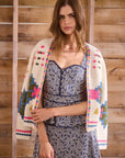Woman wearing a colorful crochet cardigan over a patterned dress against a wooden background