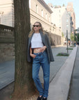 Woman standing against a tree on a city sidewalk wearing sunglasses, a white crop top, blue jeans, and a gray coat.