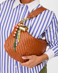 Person holding a brown woven handbag with decorative elements against a blue and white striped shirt background.