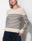 A woman wearing a cream and navy blue horizontally striped knit sweater with dark navy wide-leg trousers, standing against a plain light gray wall.