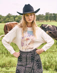 Woman in a cowboy hat and bandana-patterned skirt standing in a field with cows.