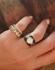 Hand wearing two gold rings with gemstones against a brown background