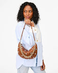A woman wearing a light blue and white color-block shirt and jeans carries a brown crescent-shaped leather shoulder bag decorated with gold-tone chain and silver studs, set against a plain white background.