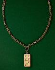 A gold-tone chain necklace with a rectangular domino pendant, displayed against a solid dark green background.