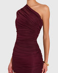 A woman wearing a ruched, one-shoulder mini dress in deep burgundy against a light gray studio background.