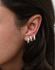 A close-up of a woman's ear wearing three small hoop earrings with clear gemstones, shown against her dark hair and skin.