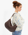 A woman in a cream knit sweater with blue speckles and jeans is shown from the side, carrying a large brown woven crescent shoulder bag with gold-tone hardware against a plain white background.
