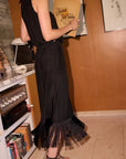 A woman wearing a sleeveless black midi set with a fringe hem stands in a room
