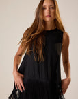 Woman wearing a black sleeveless top with fringe details against a plain background