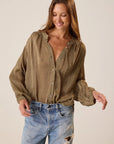 A woman wearing a brown and beige patterned long-sleeve blouse with a split neckline and button front, paired with light-wash distressed jeans, against a plain beige studio background.