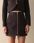 A model wearing a dark brown cropped jacket and matching high-waisted mini skirt with contrast light stitching and a front zipper, set against a plain beige background.