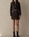 A model stands against a plain beige studio backdrop wearing a dark brown cropped zip-front jacket and matching mini skirt set, paired with dark ankle boots.