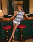 Woman in a blue and white matching set standing between two red stools in a bar setting.