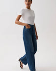 A woman modeling a white short-sleeve top and high-waisted, wide-leg blue denim jeans against a plain light gray studio background.